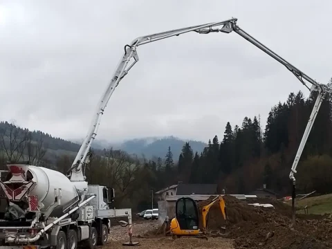 Concrete pump with extended boom and a truck mixer on a construction site, wooded hills in the background