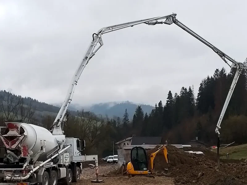 Concrete pump with extended boom and a truck mixer on a construction site, wooded hills in the background