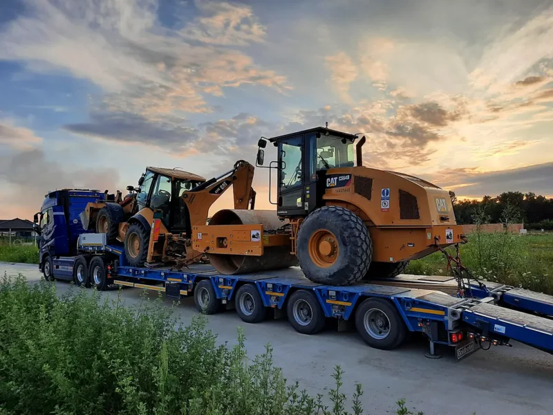 Ťahač s podvalníkom prepravuje cestný valec CAT a rýpadlo-nakladač CAT pri západe slnka