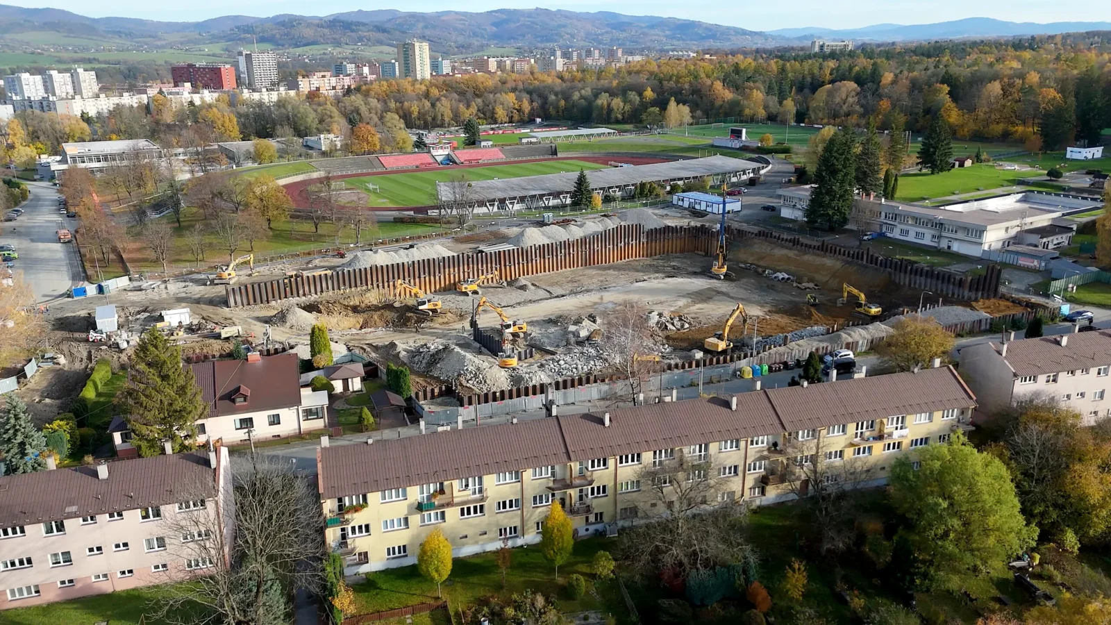 Panoramatický letecký pohľad na stavebnú jamu so štetovnicovou stenou medzi bytovými domami a štadiónom
