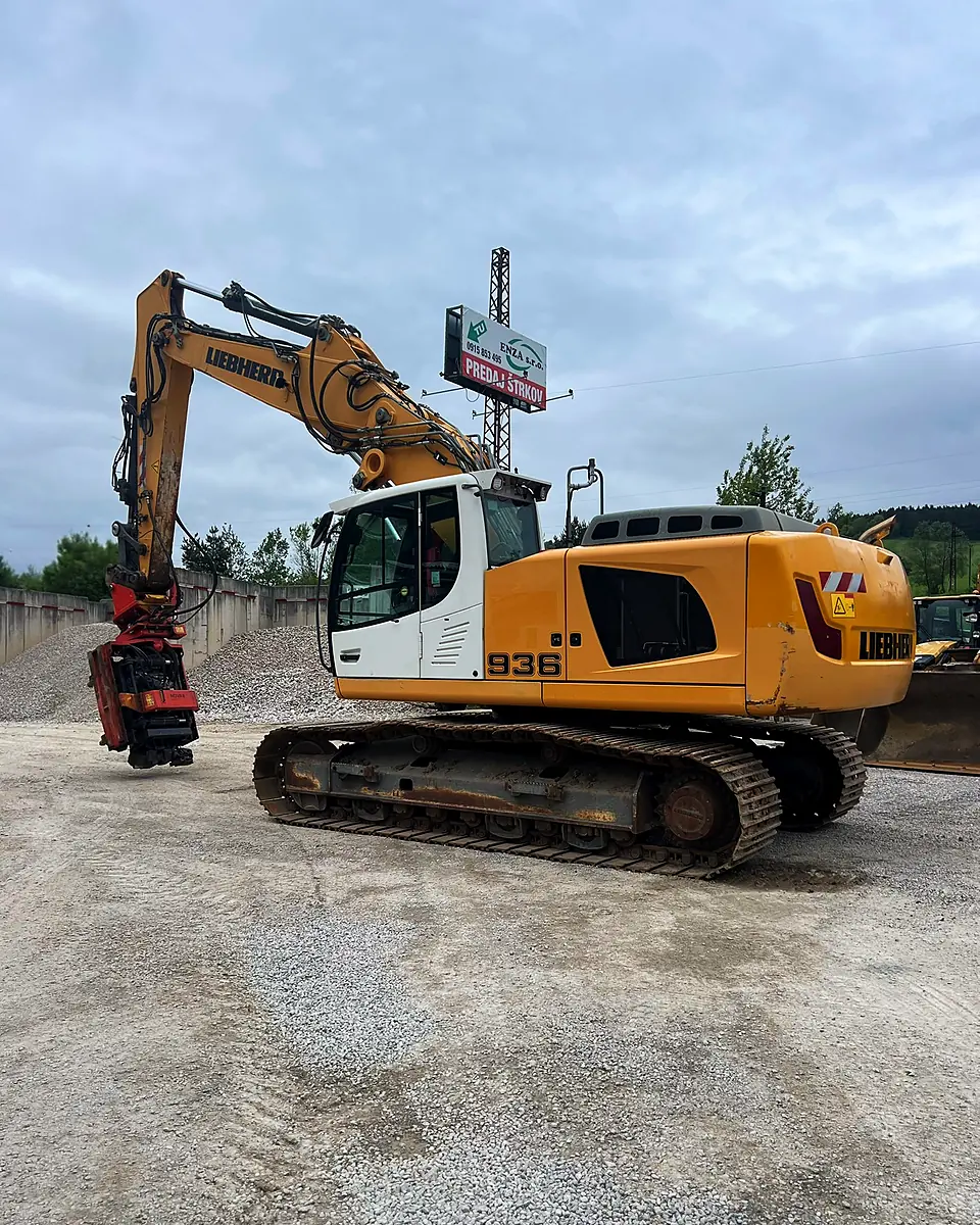 Liebherr excavator with a MOVAX vibratory pile driver driving sheet piles on a gravel plateau beside a building