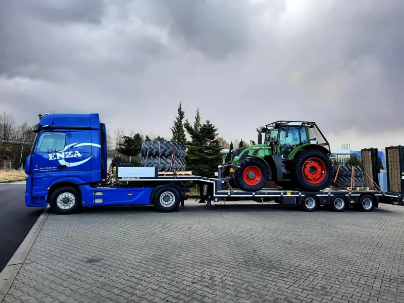 Modrý ťahač ENZA s podvalníkom prepravuje zelený traktor